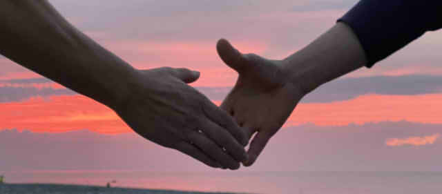 夕日をバックに手をつなごうとしている男性二人の手の画像