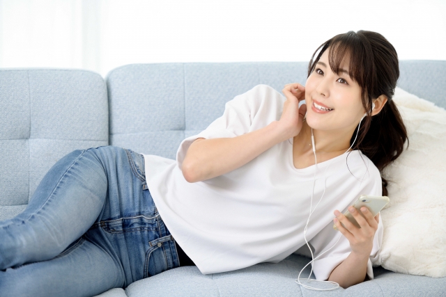 ソファで寝そべってイヤホンをつける女性の画像