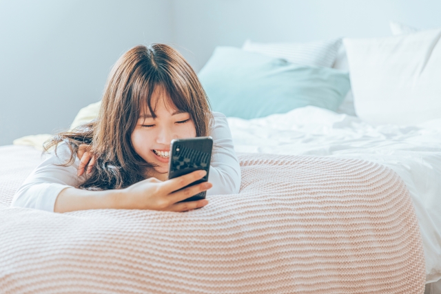 ベッドでスマホを見ながら楽しむ女性の画像