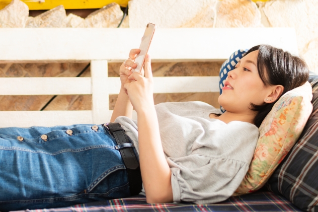 ソファーで寝ながらスマホを触る女性の画像